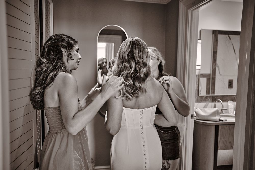 Black and white photo of bridesmaids helping the bride with her wedding dress buttons.
