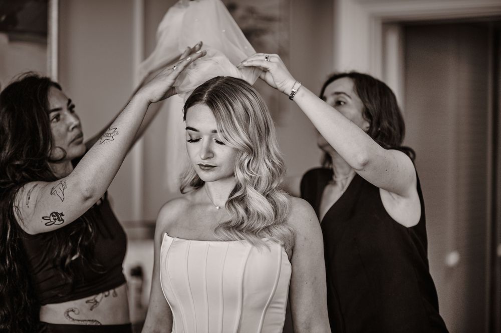 Two assistants placing a wedding veil on a blonde bride during bridal preparations in black and white.