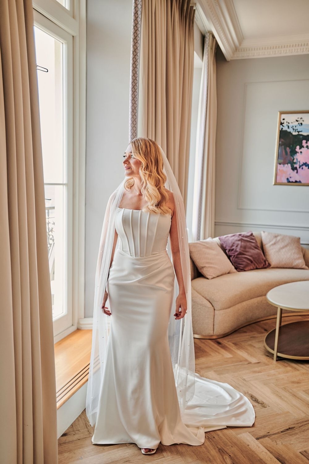 Elegant bride in strapless white gown with long veil standing by tall windows in luxury hotel suite.