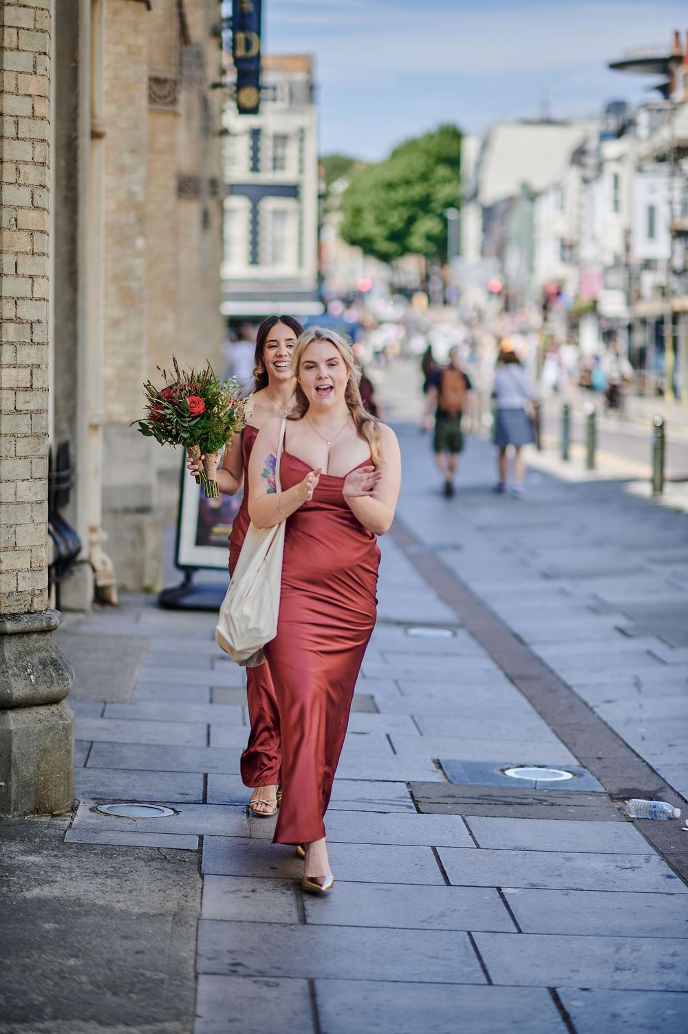 Two women in red satin dresses walk down a sunny urban street, one holding a bouquet of flowers.
