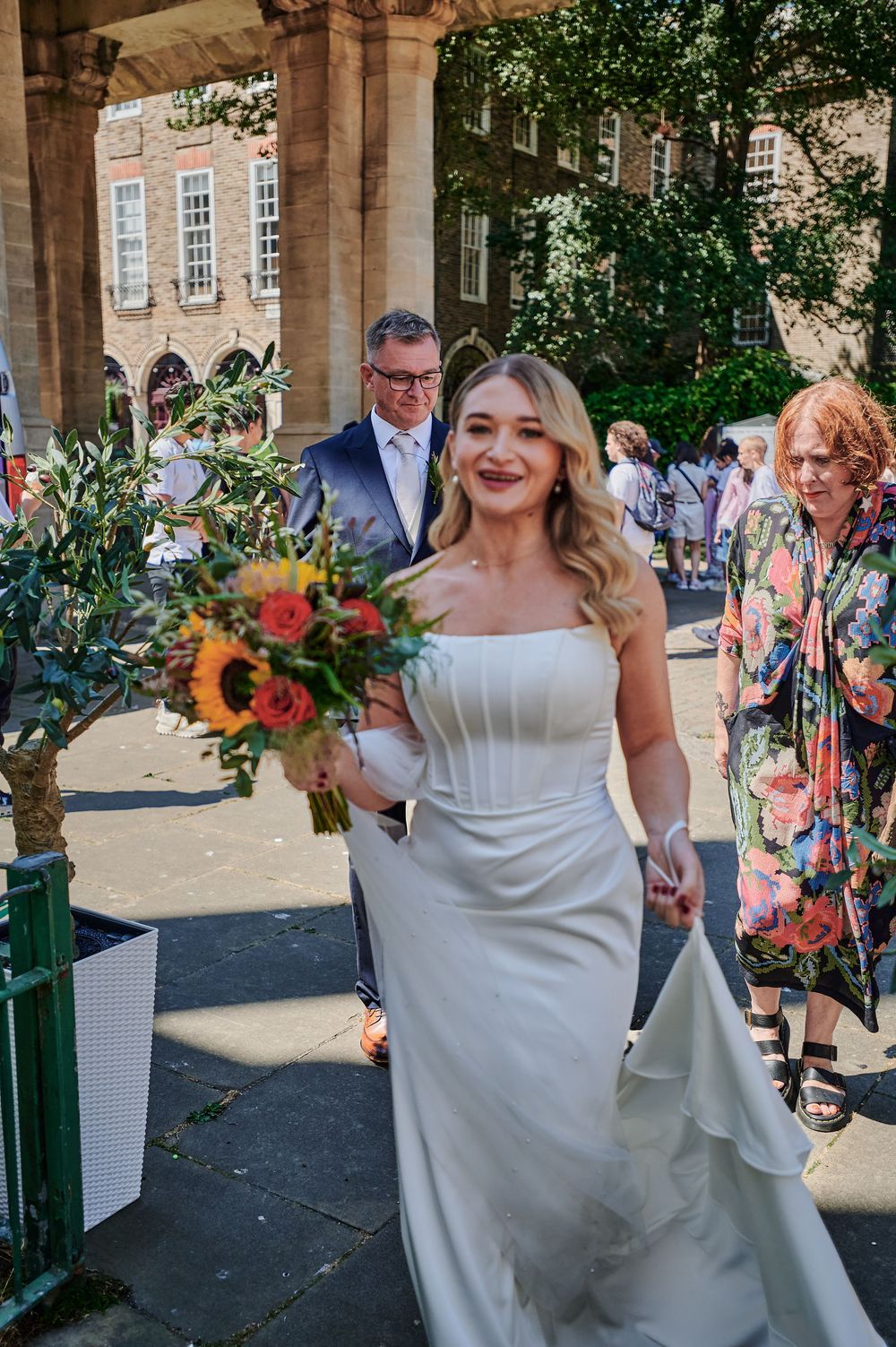 Smiling bride in strapless white gown holding vibrant sunflower bouquet walks outside historic brick venue with guests.