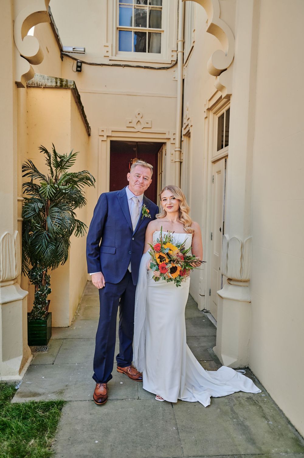 Bride holding sunflower bouquet and groom in navy suit pose outside elegant cream building on their wedding day.