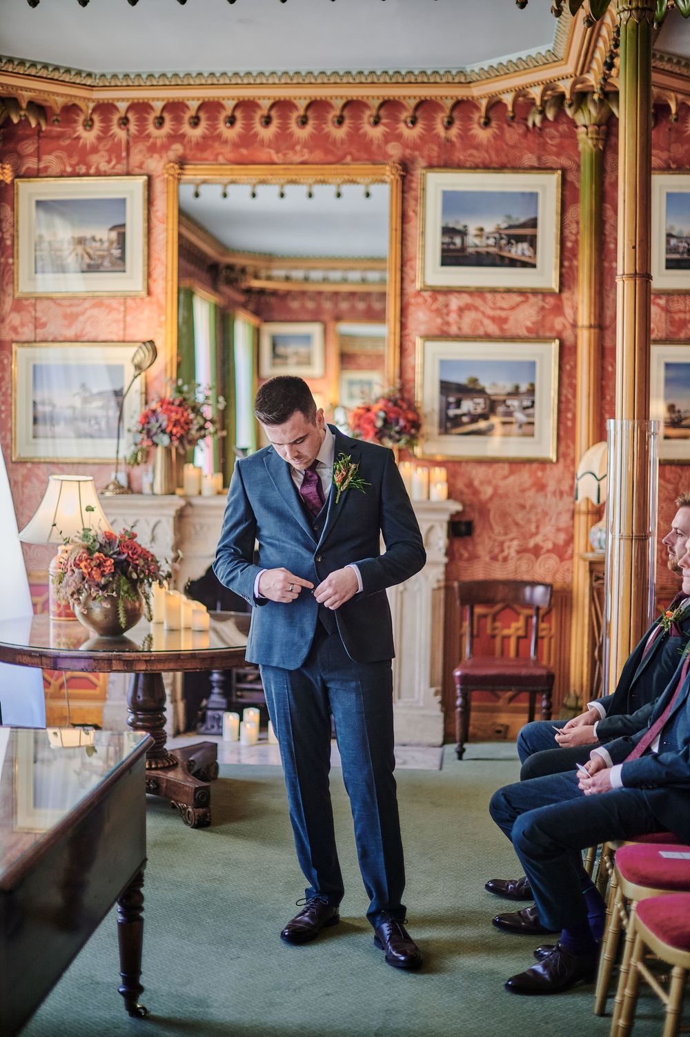 Groom in navy suit adjusting jacket button in ornate red-walled room before wedding ceremony.