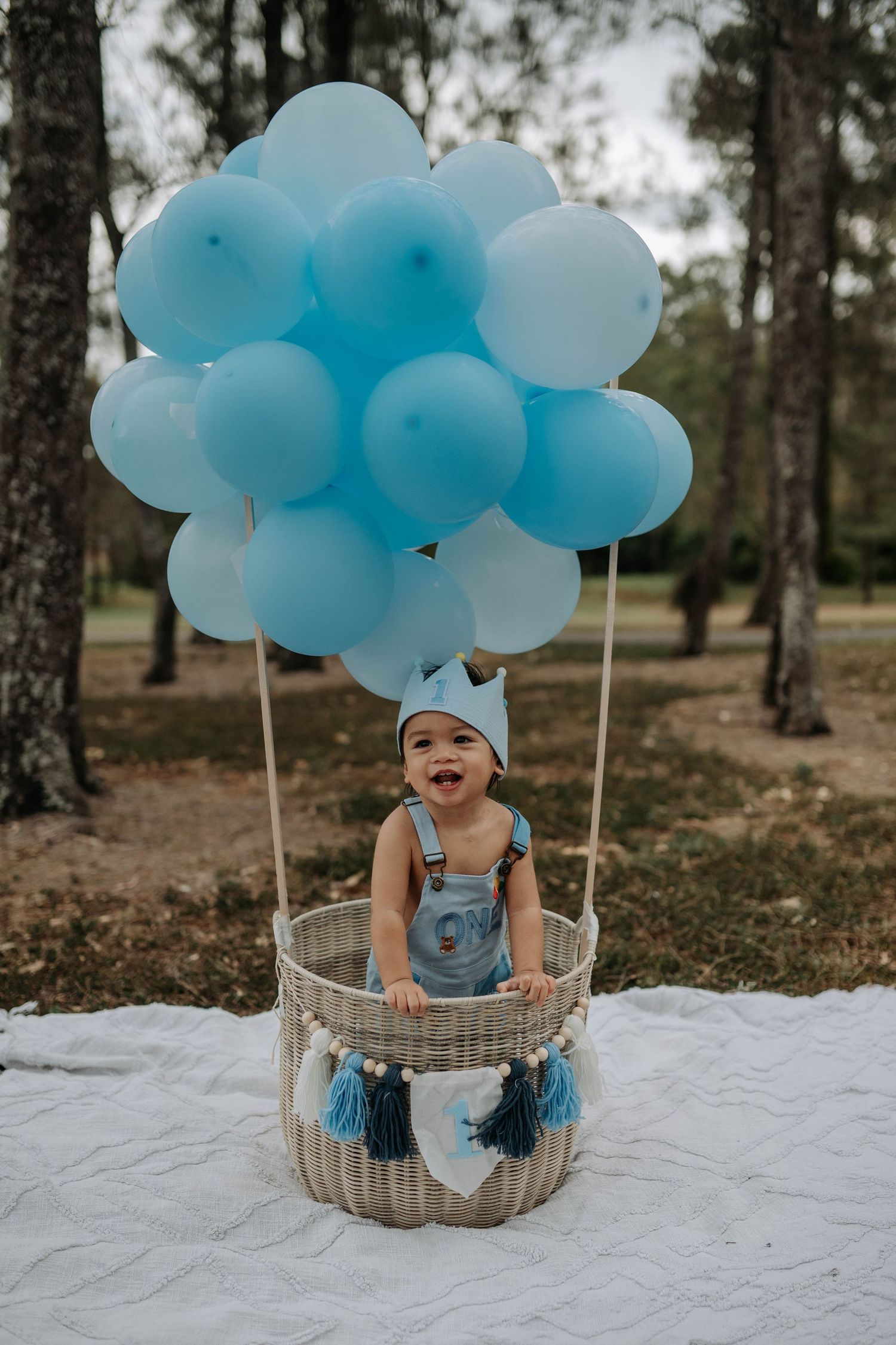 Baby boy in denim overalls sits in a wicker basket decorated with blue balloons for a first birthday photo shoot.
