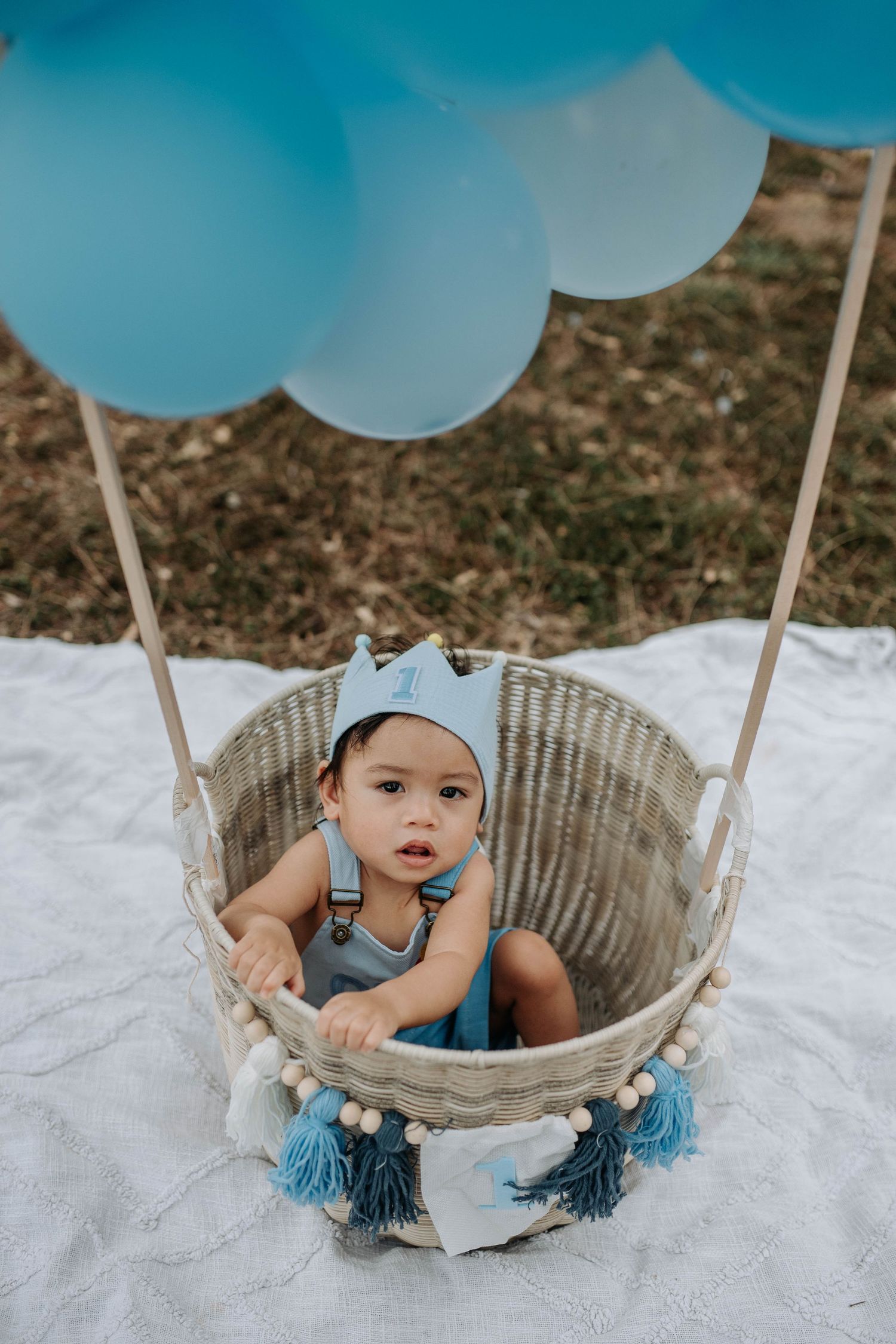 Baby boy in blue outfit sits in wicker basket with blue balloons, celebrating his first birthday outdoors.