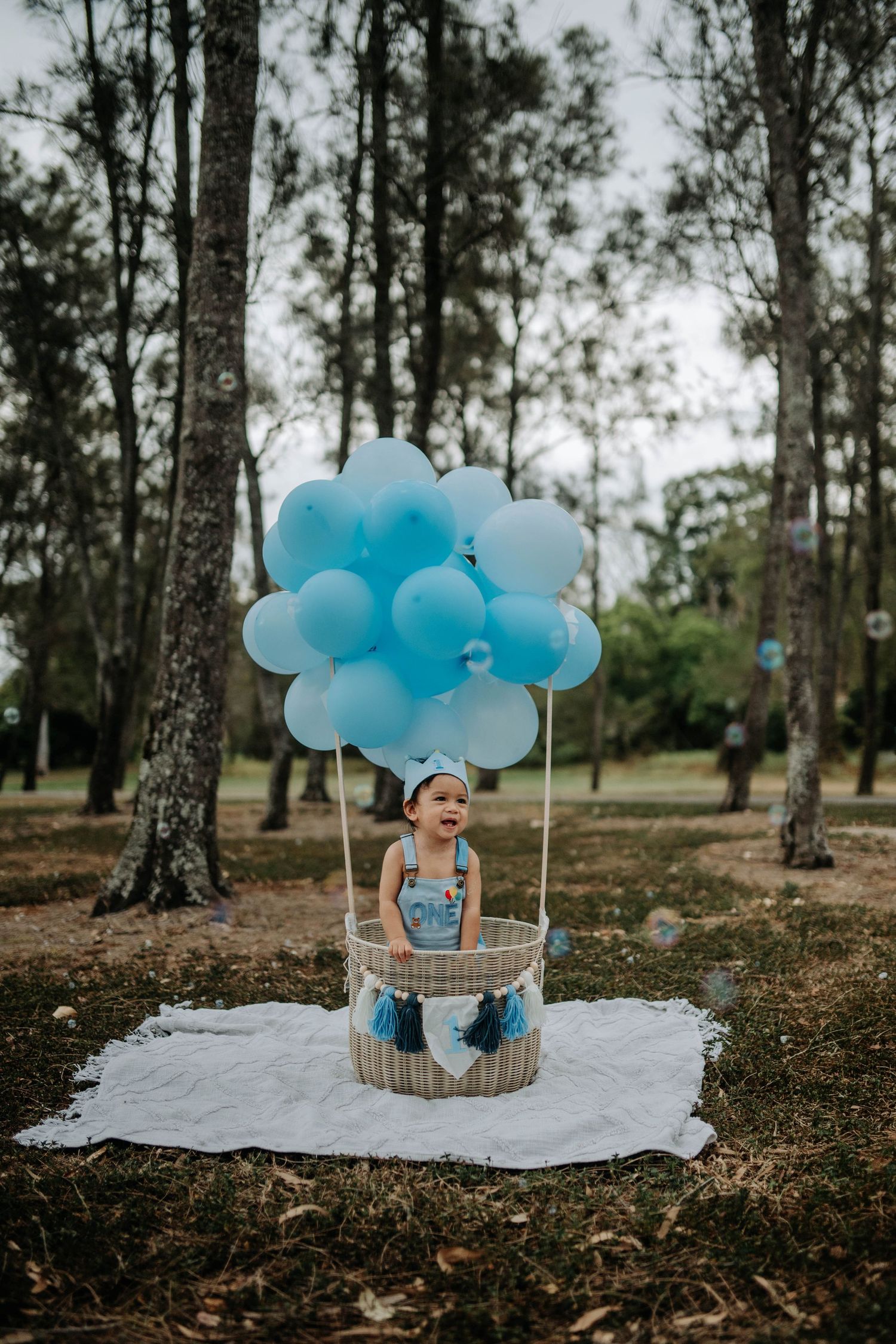 Toddler sitting in a wicker basket with blue balloons arranged as a hot air balloon in a park.