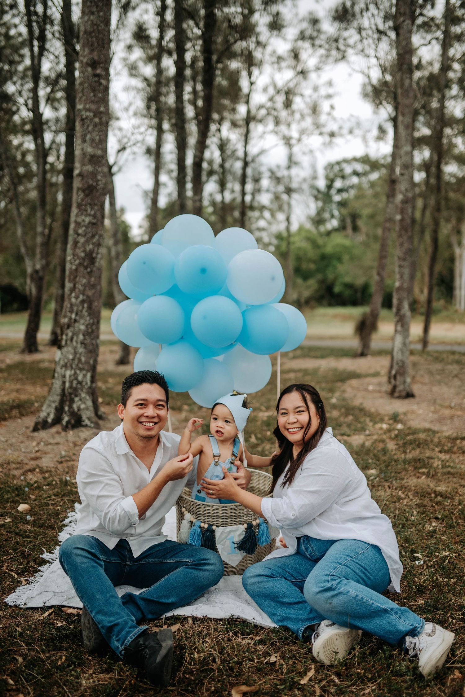 Happy family with baby celebrating in a park, surrounded by blue balloons on a picnic blanket among trees.
