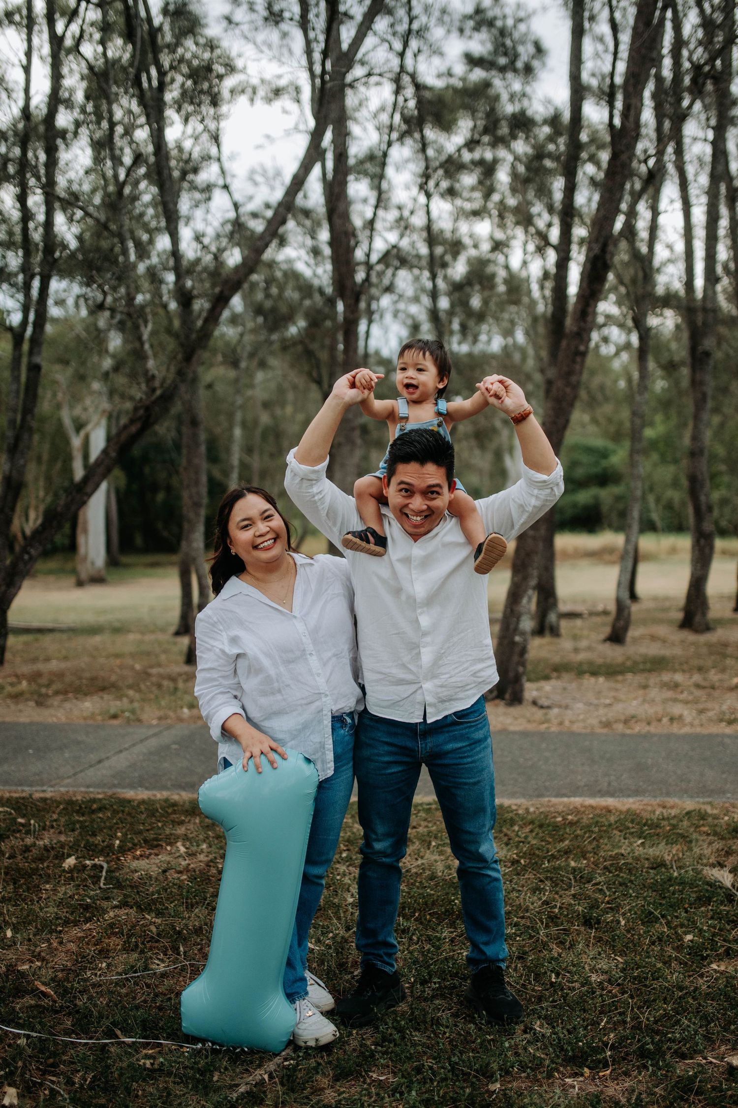 A happy Asian family poses outdoors, parents in white shirts and jeans, child on dad's shoulders, holding a teal number one balloon.