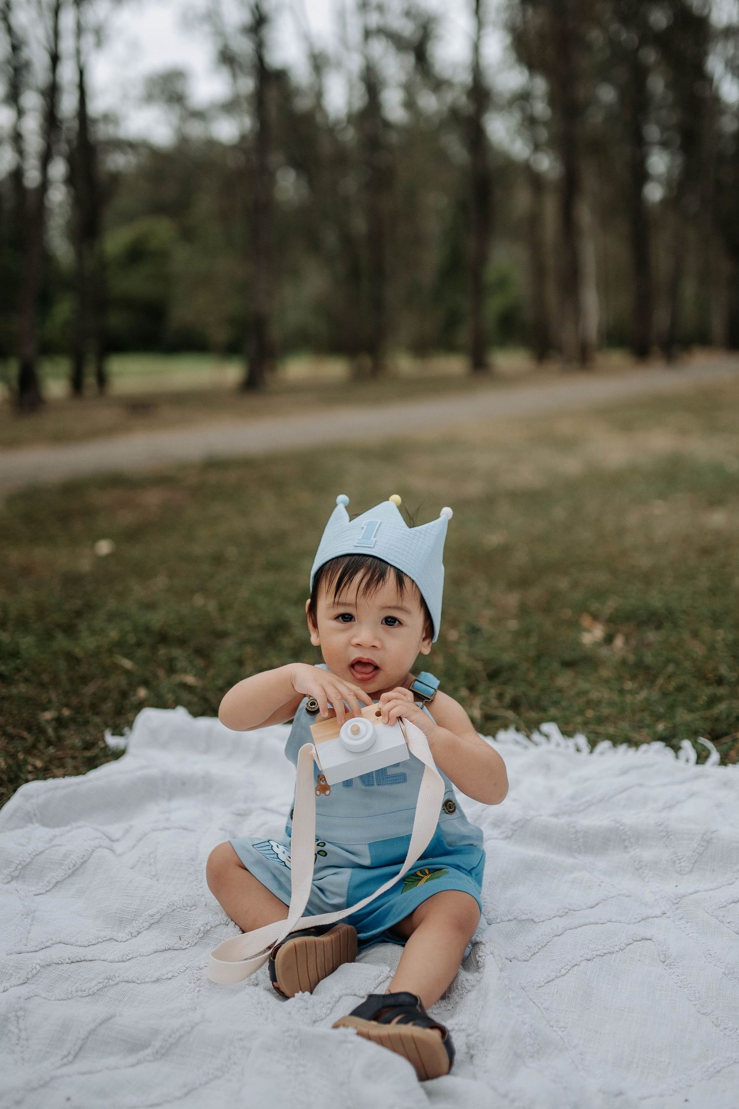 A baby boy wearing a blue crown hat sits on a white blanket in a park, holding a toy camera.