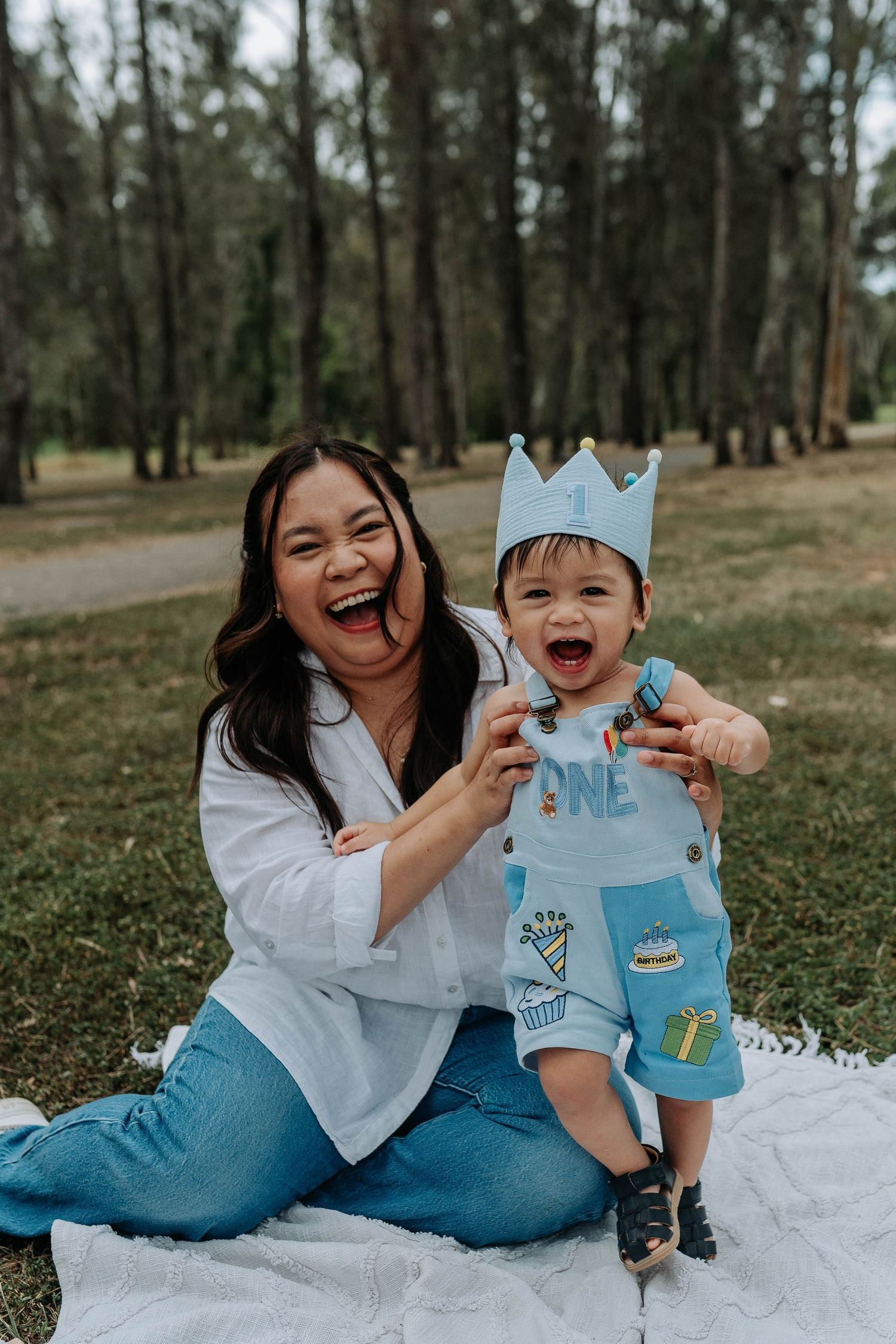 A laughing mother sits on a blanket in a park with her baby boy wearing a blue crown and ONE birthday outfit.