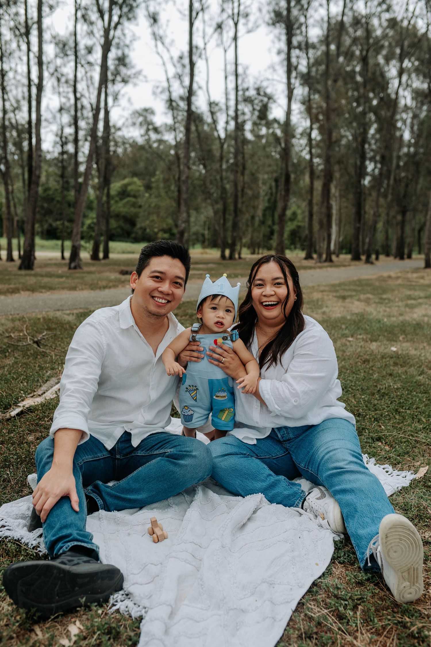 Happy Asian family sitting on a blanket in a park, parents in white shirts and jeans with their baby wearing a blue crown.