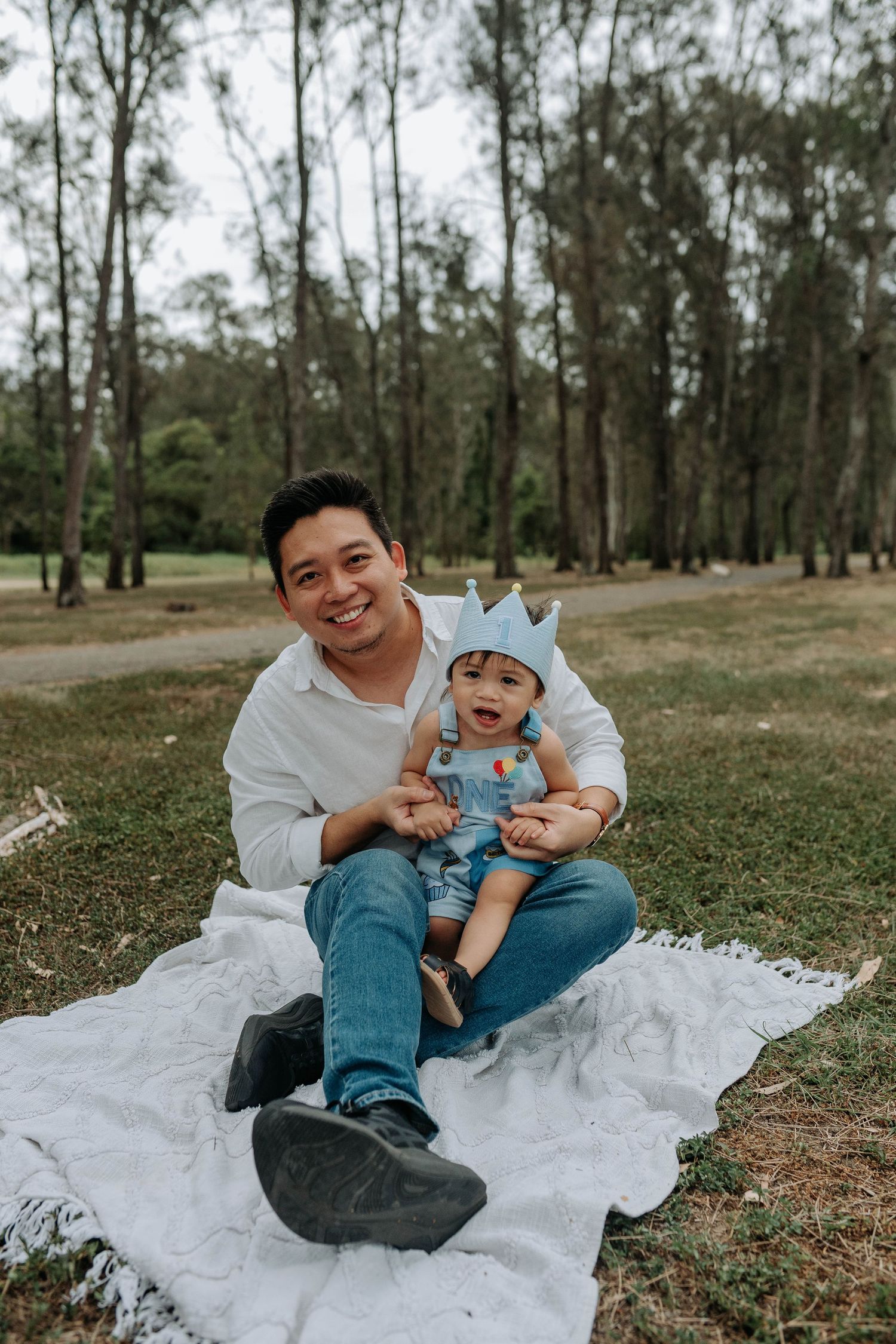 A smiling man sits on a white blanket in a park holding a laughing baby wearing a blue crown hat.