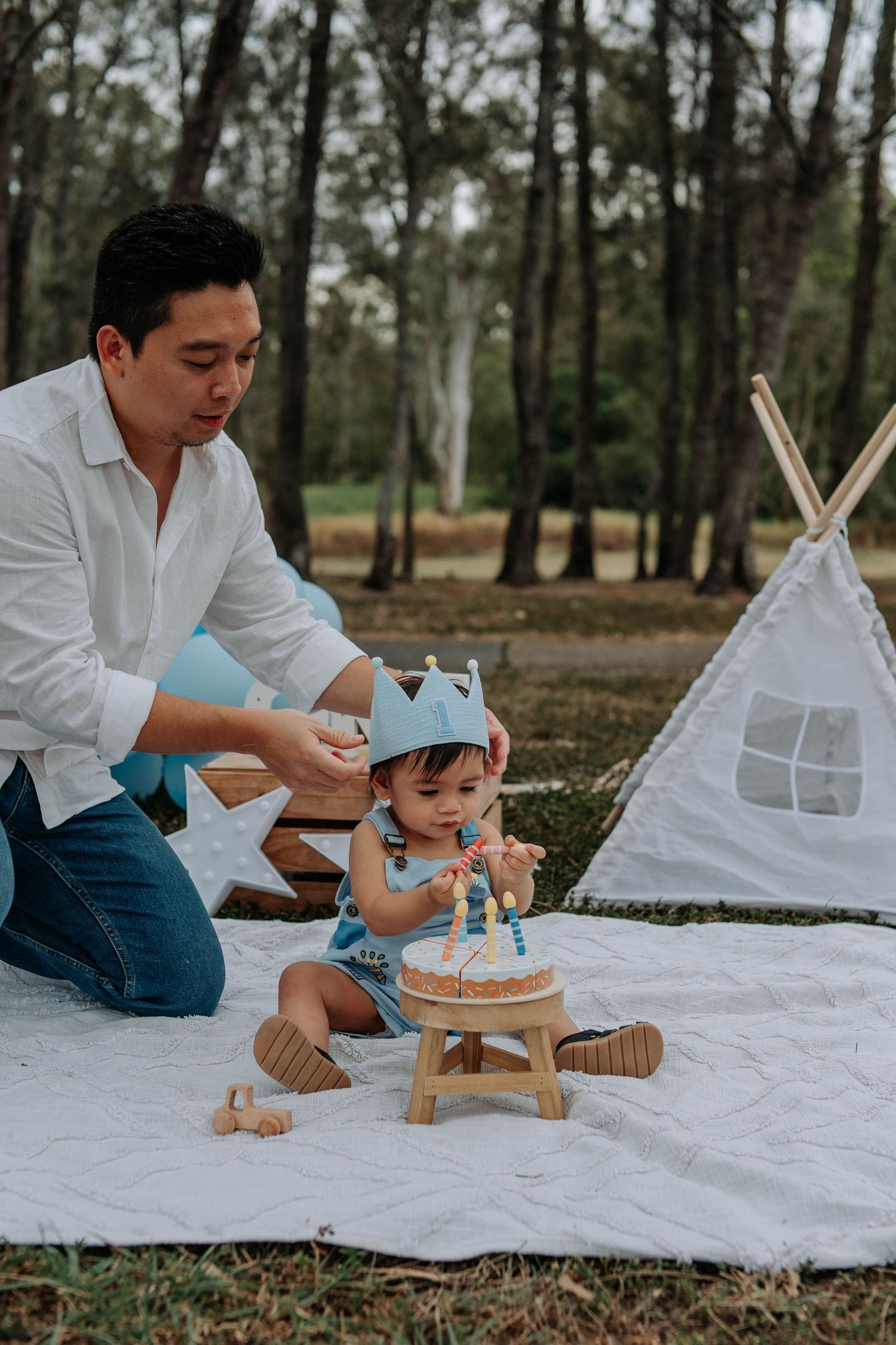 Father helps toddler with birthday crown beside small cake outdoors on white blanket near teepee in forest setting.