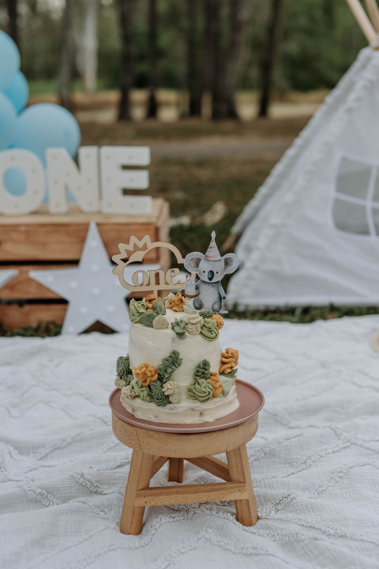 First birthday cake with koala topper and floral decorations on wooden stool at outdoor ONE party.