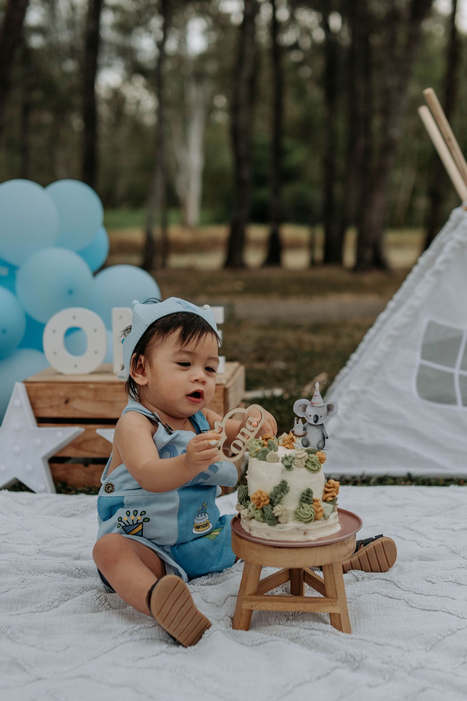 Baby boy in blue romper reaching for first birthday cake outdoors, with balloons and teepee backdrop.
