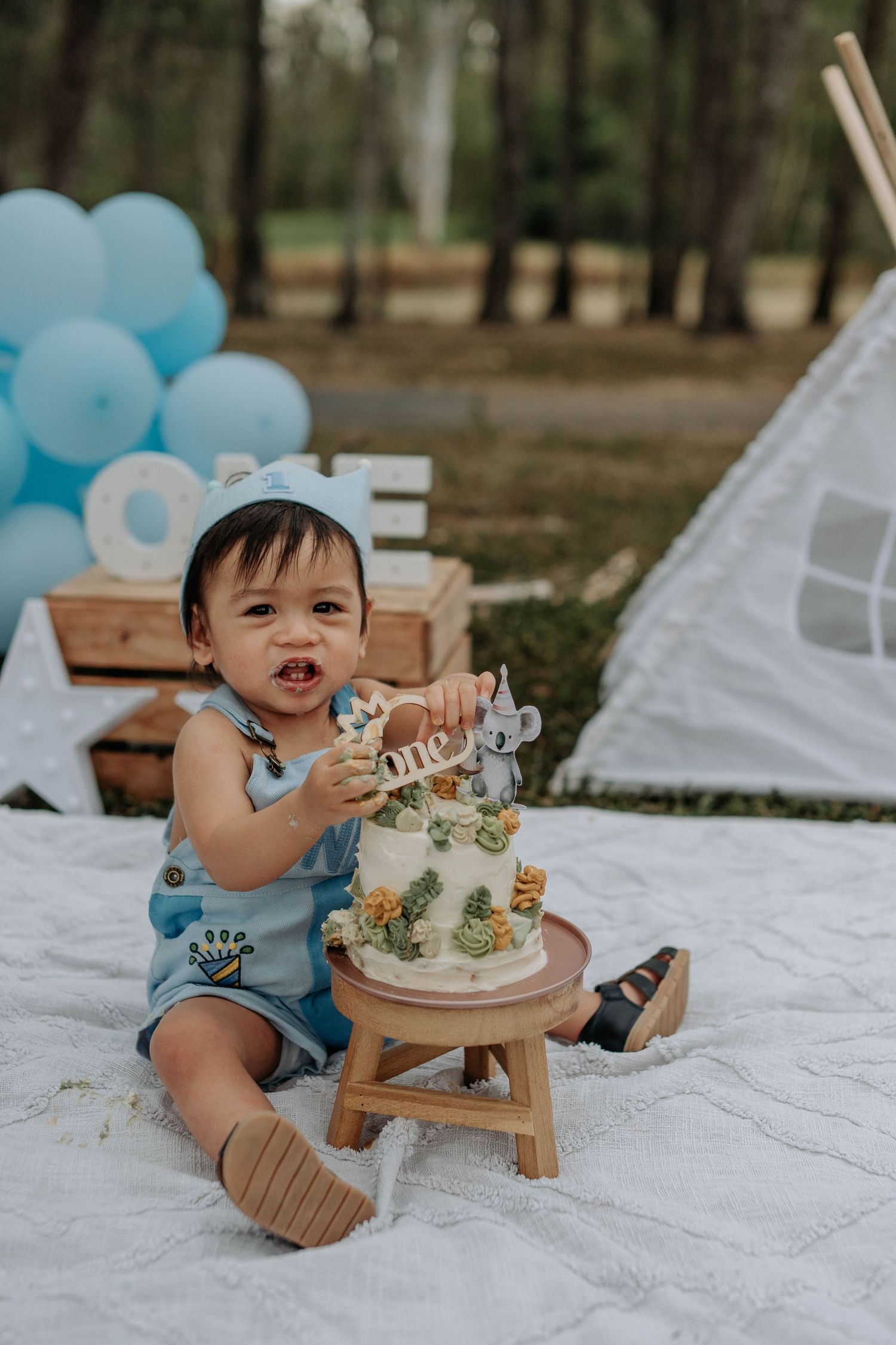 Baby boy in blue romper smashes first birthday cake outdoors with balloon decorations and teepee backdrop.