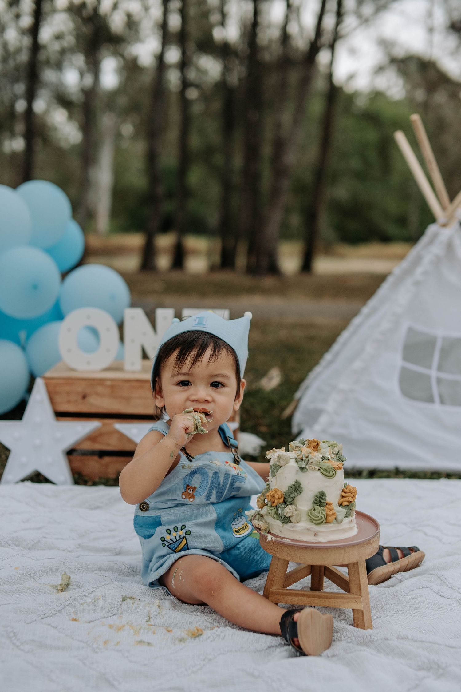 Baby boy in blue romper eating first birthday cake outdoors with balloons and ONE sign backdrop.