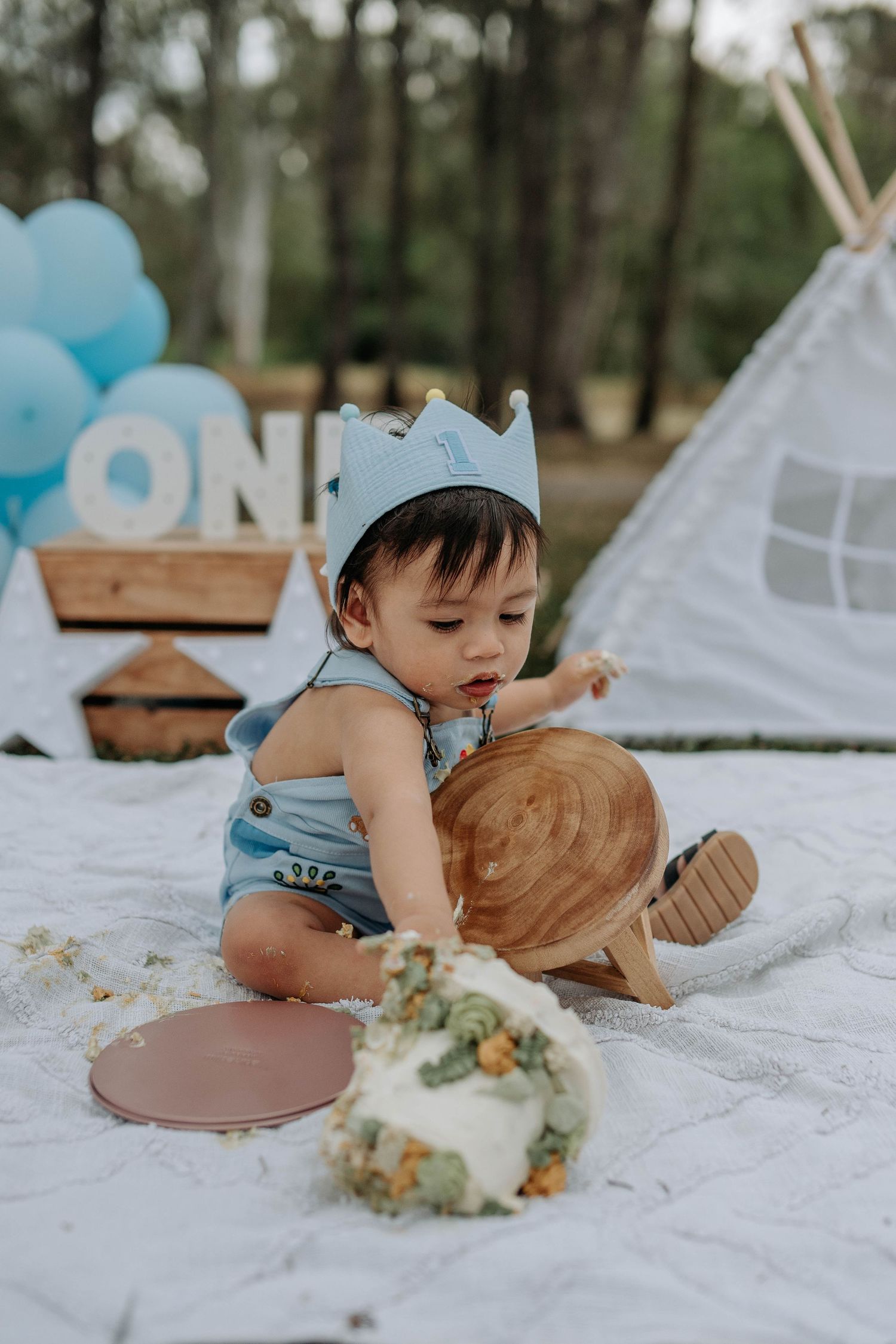 Baby boy in blue romper and crown smashes first birthday cake outdoors near teepee and balloons.