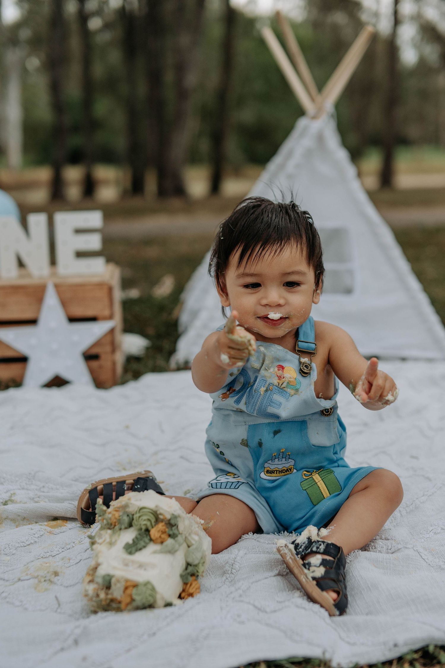 Baby boy in blue overalls smashes first birthday cake outdoors near a teepee and ONE sign.