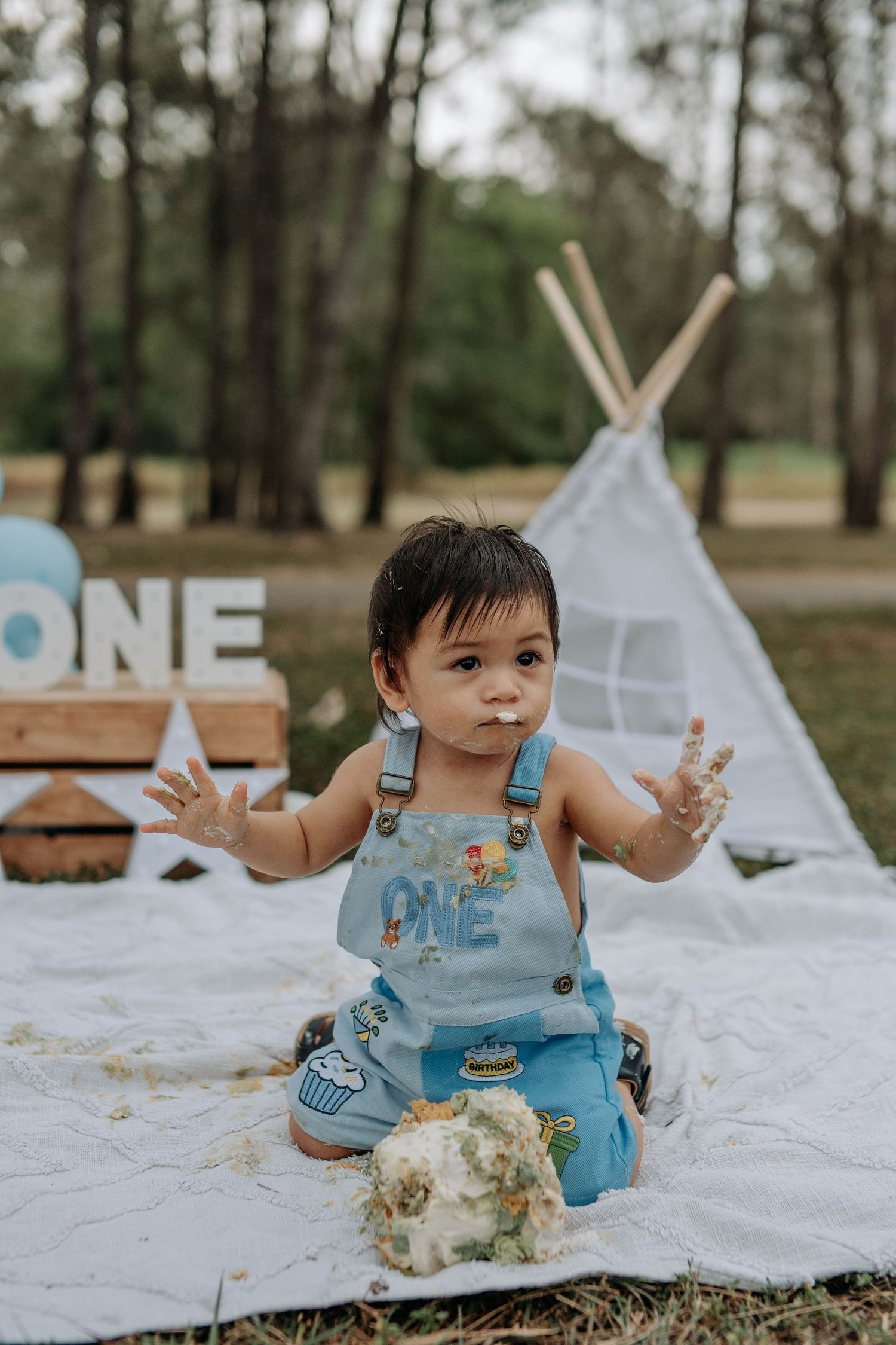 A baby boy in blue ONE overalls celebrates his first birthday with a smashed cake outdoors near a white teepee.