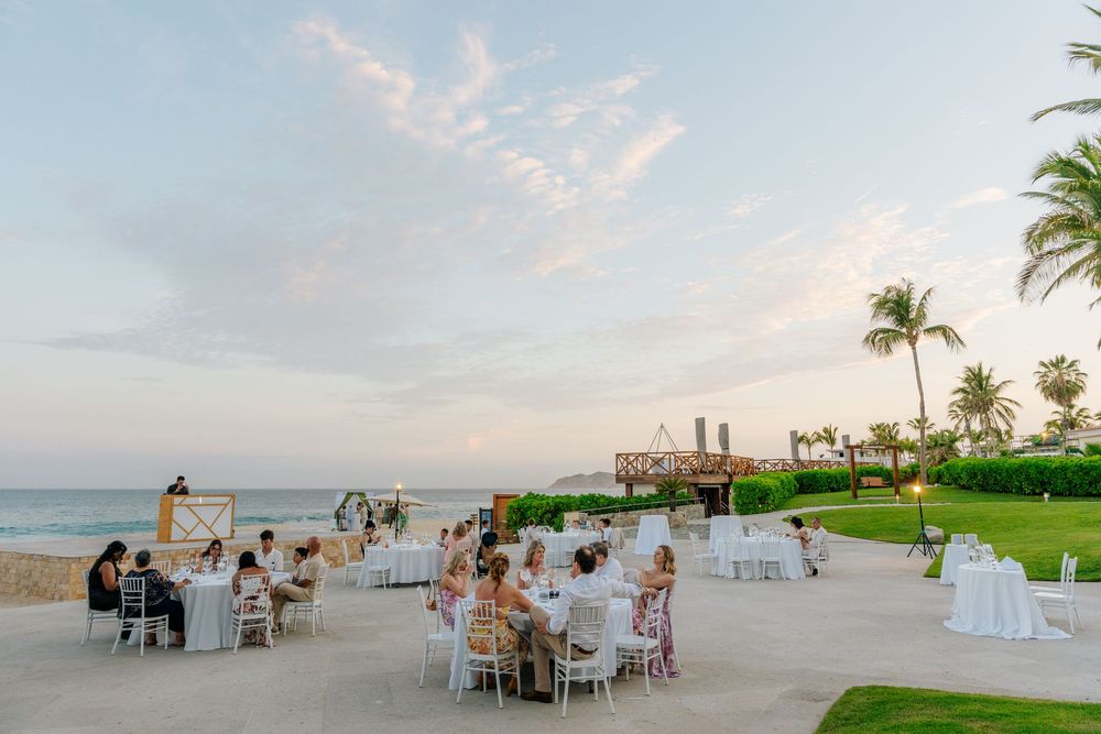 Guests dine at white-clothed tables on a beachfront terrace at sunset, surrounded by palm trees and ocean views at a tropical resort.