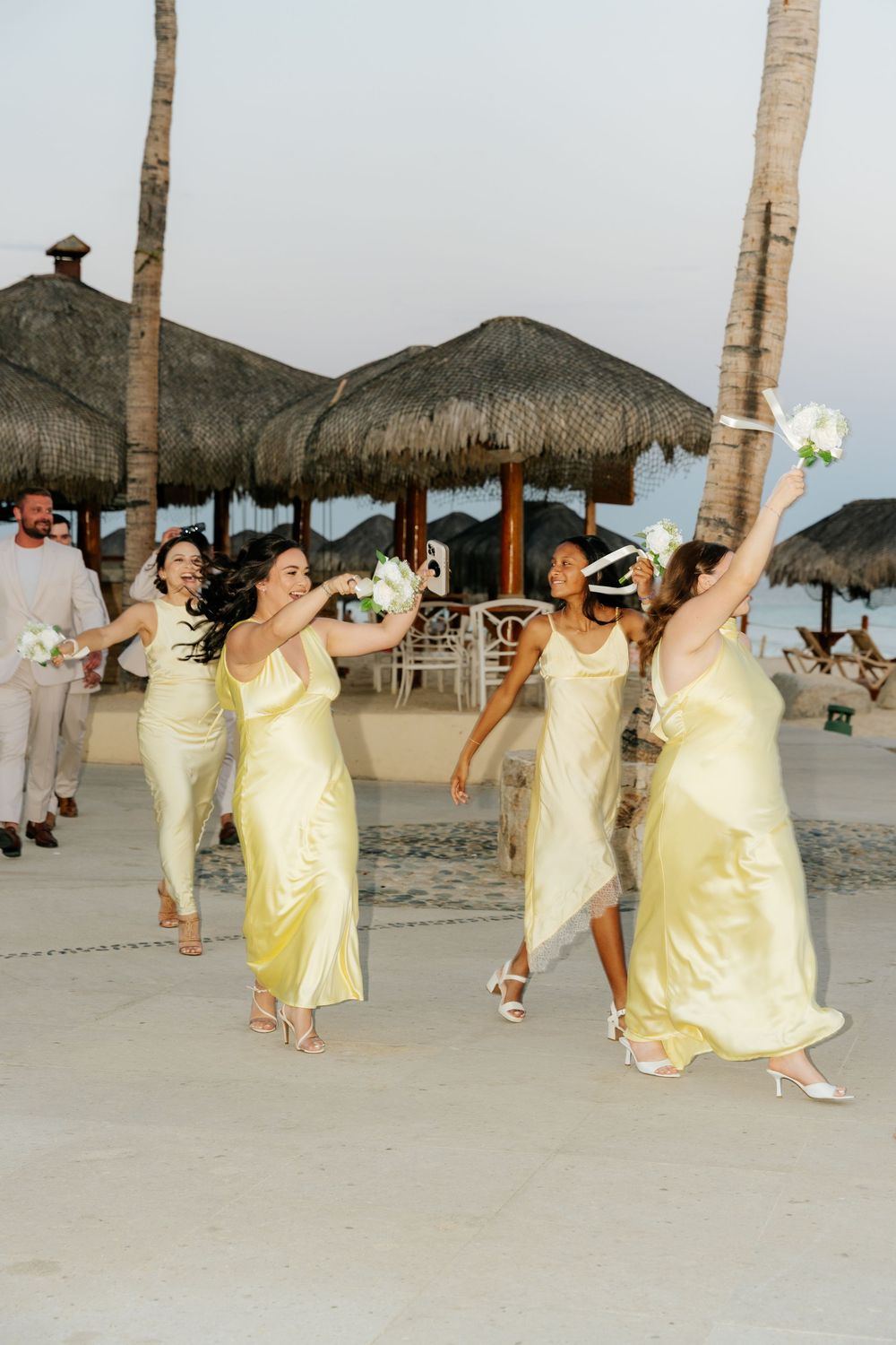 Bridesmaids in pale yellow satin dresses dance with bouquets at a beachfront wedding near thatched palapas and palm trees.