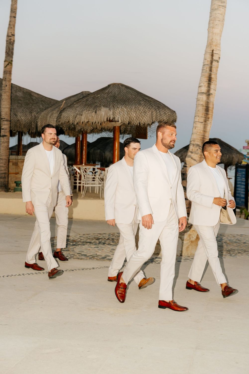 Four men in cream linen suits and brown leather shoes walk along a sandy beach path past thatched palapa huts at sunset.