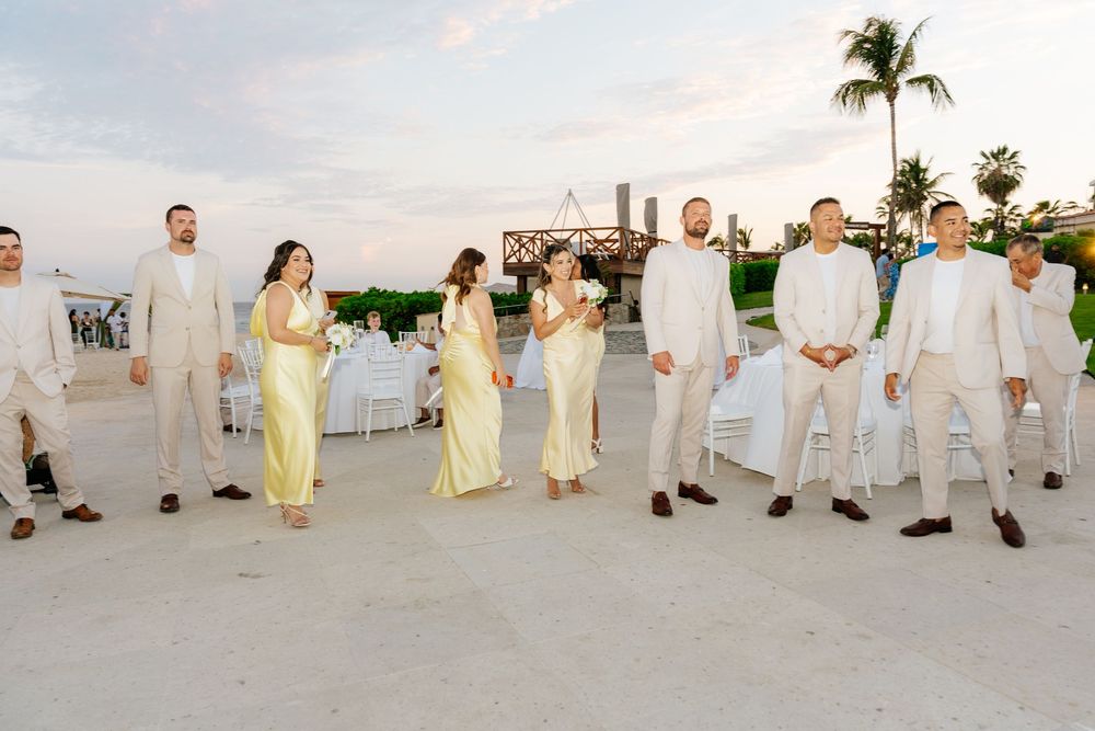 Wedding party in tan suits and yellow dresses stands on a beachfront terrace at sunset with palm trees and white chairs in the background.