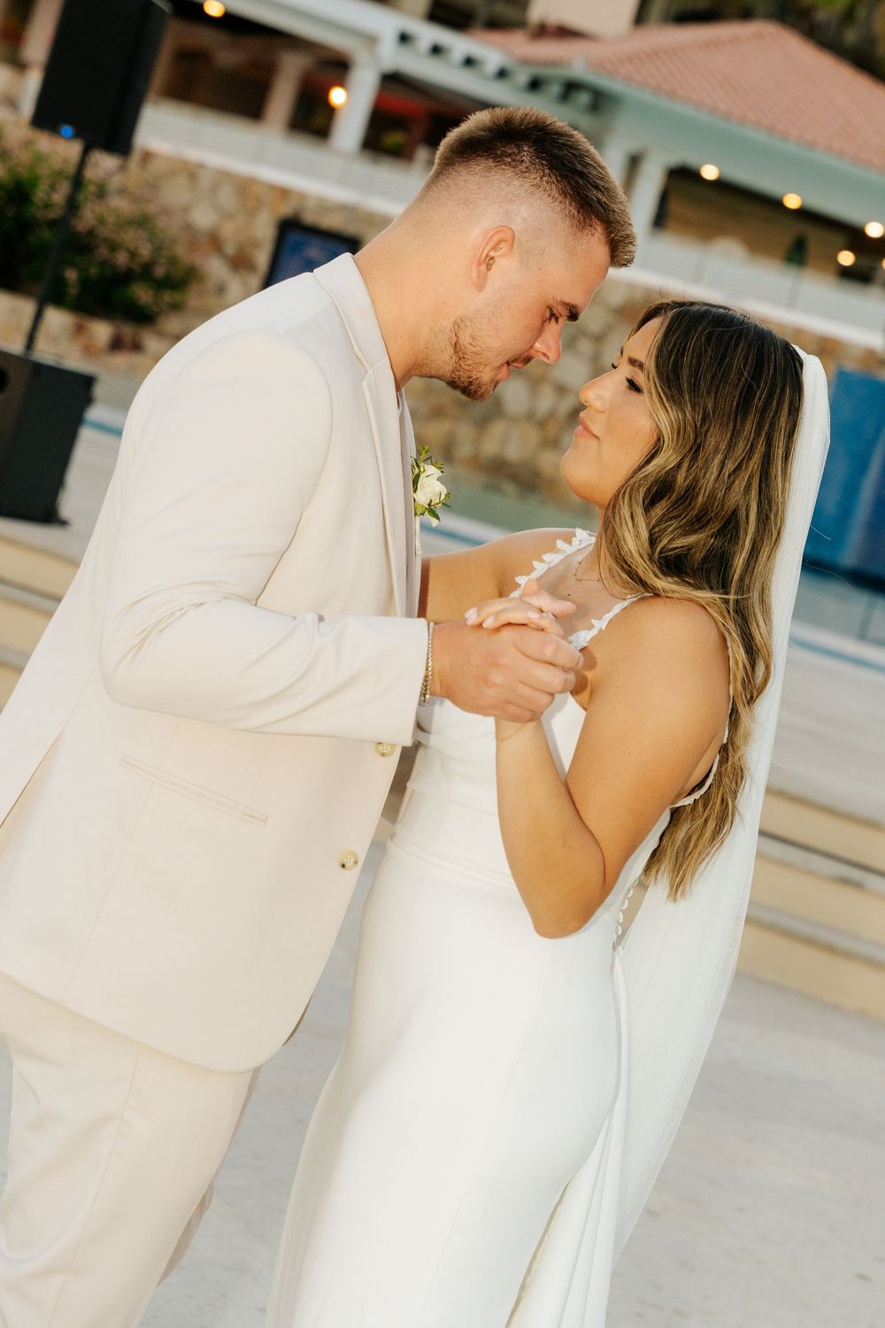 A groom in a cream suit dances closely with a bride in a white gown and long veil at an outdoor beach resort wedding.