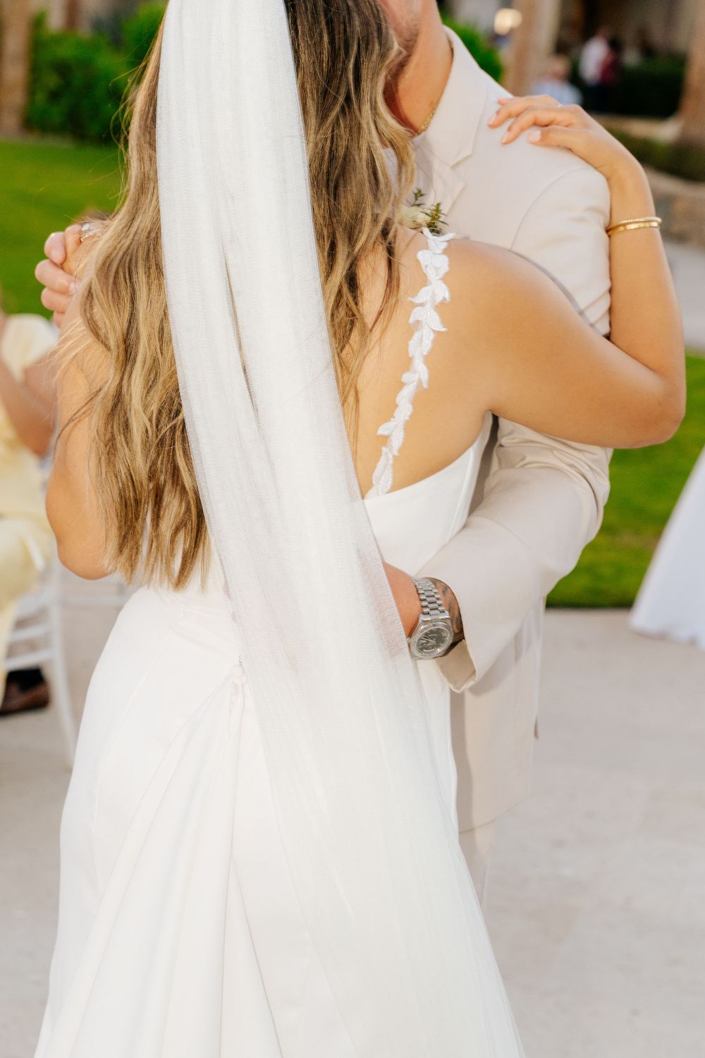 Bride in a white gown with lace strap and long veil embraces a groom in a beige suit during a romantic outdoor wedding dance.