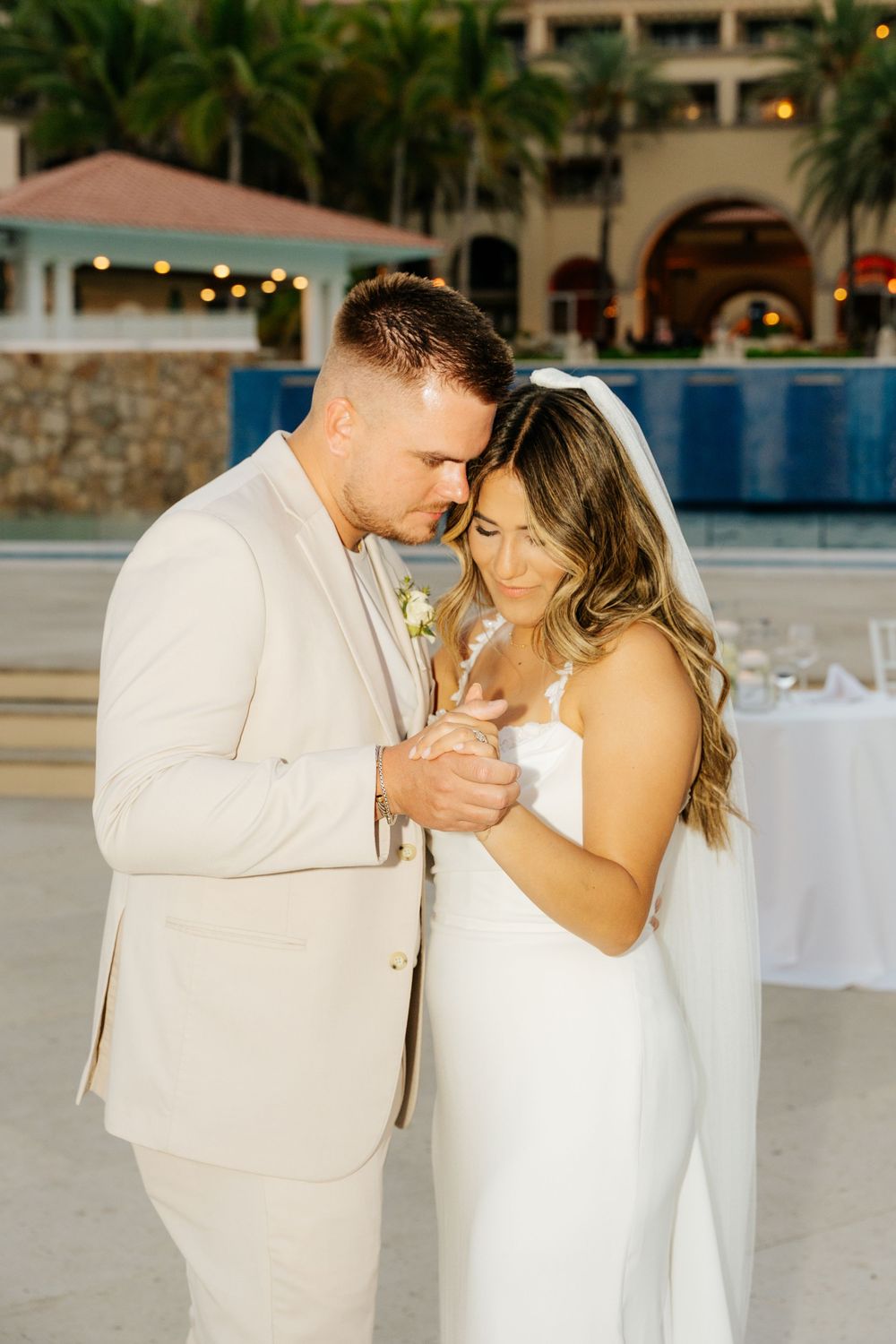 Bride in a white gown and veil shares a tender moment with groom in a cream suit at an outdoor beachside wedding reception.