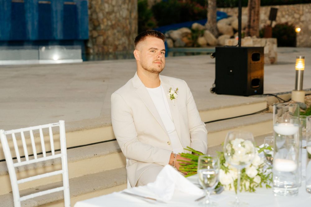 Man in a beige linen suit with a floral boutonniere sits at a white-draped wedding table with candles and flowers at an outdoor venue.