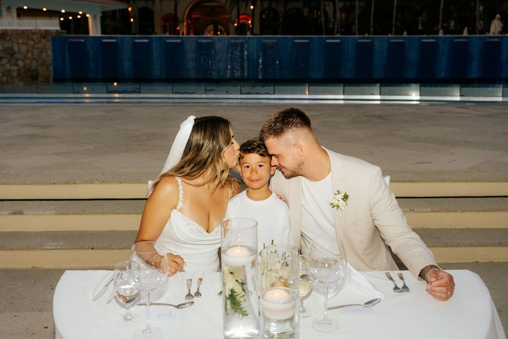 Bride in a white dress and groom in a cream suit share a kiss beside a smiling boy at a candlelit outdoor wedding dinner table.