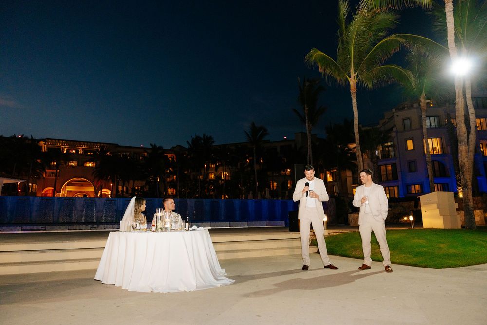 Two men in white give speeches beside a seated couple at an outdoor wedding reception table at night, with palm trees and a resort behind.