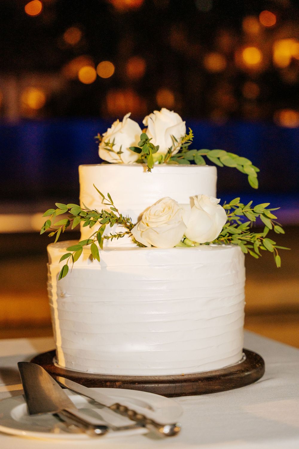 Two-tier white wedding cake topped with white roses and green foliage on a wooden stand with a cake knife and server.