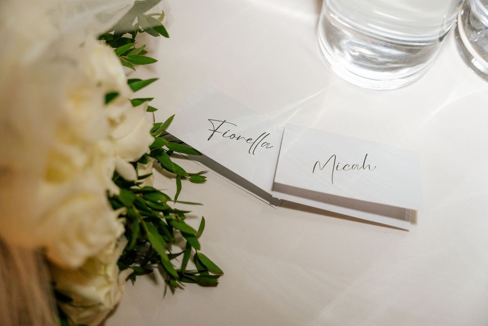 Elegant wedding place cards with handwritten names beside white roses and green foliage on a table next to a glass.