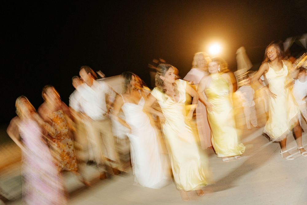 Blurred motion shot of women in flowing yellow and pastel dresses dancing outdoors at night under a glowing light.