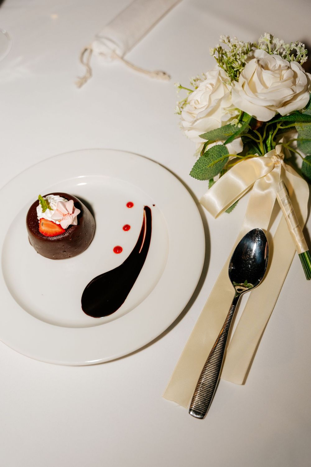 Chocolate dessert topped with strawberry and cream on a white plate beside a silver spoon and a white rose bouquet.