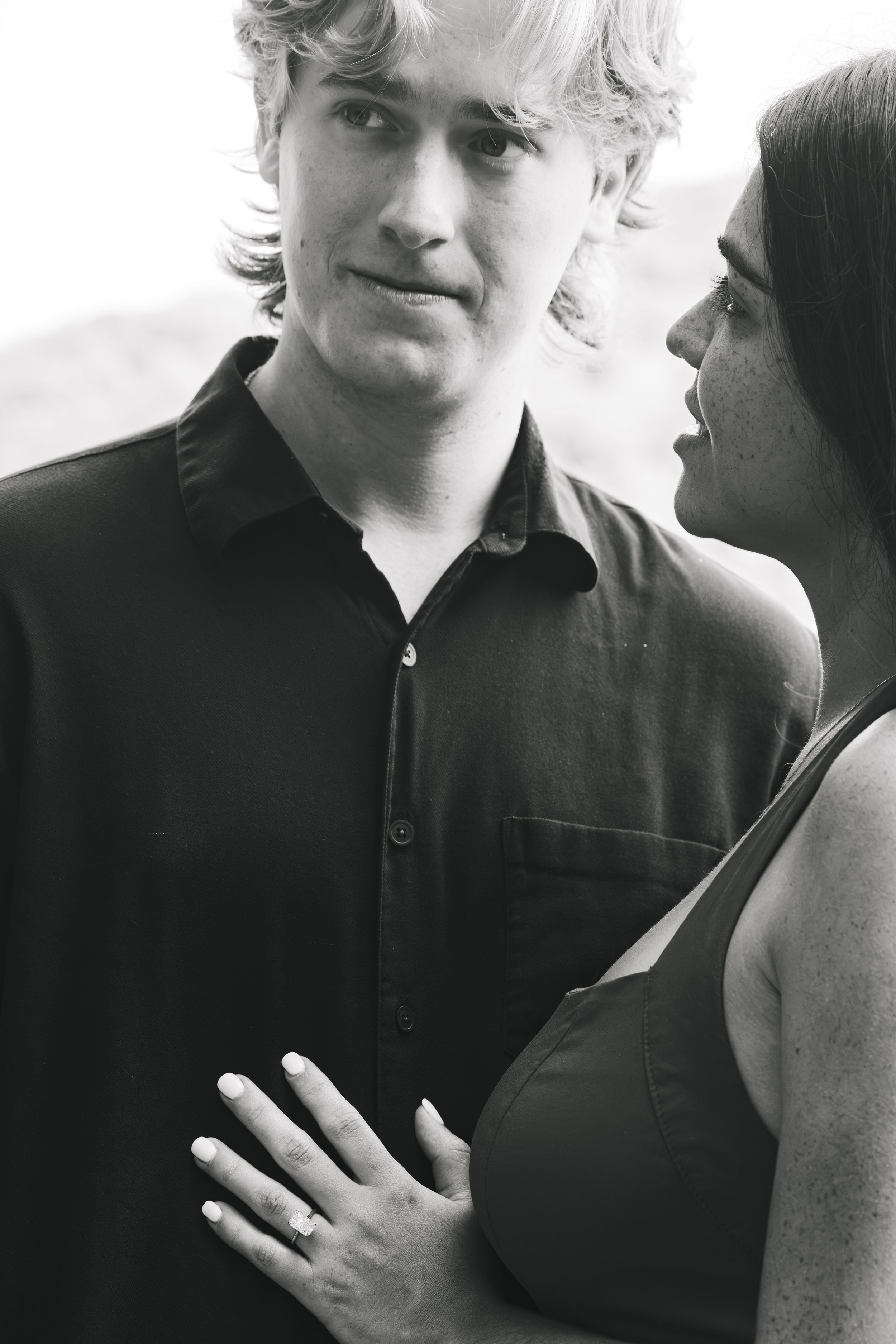 Black and white portrait of a man gazing at a woman whose hand, wearing an engagement ring, rests on his chest.