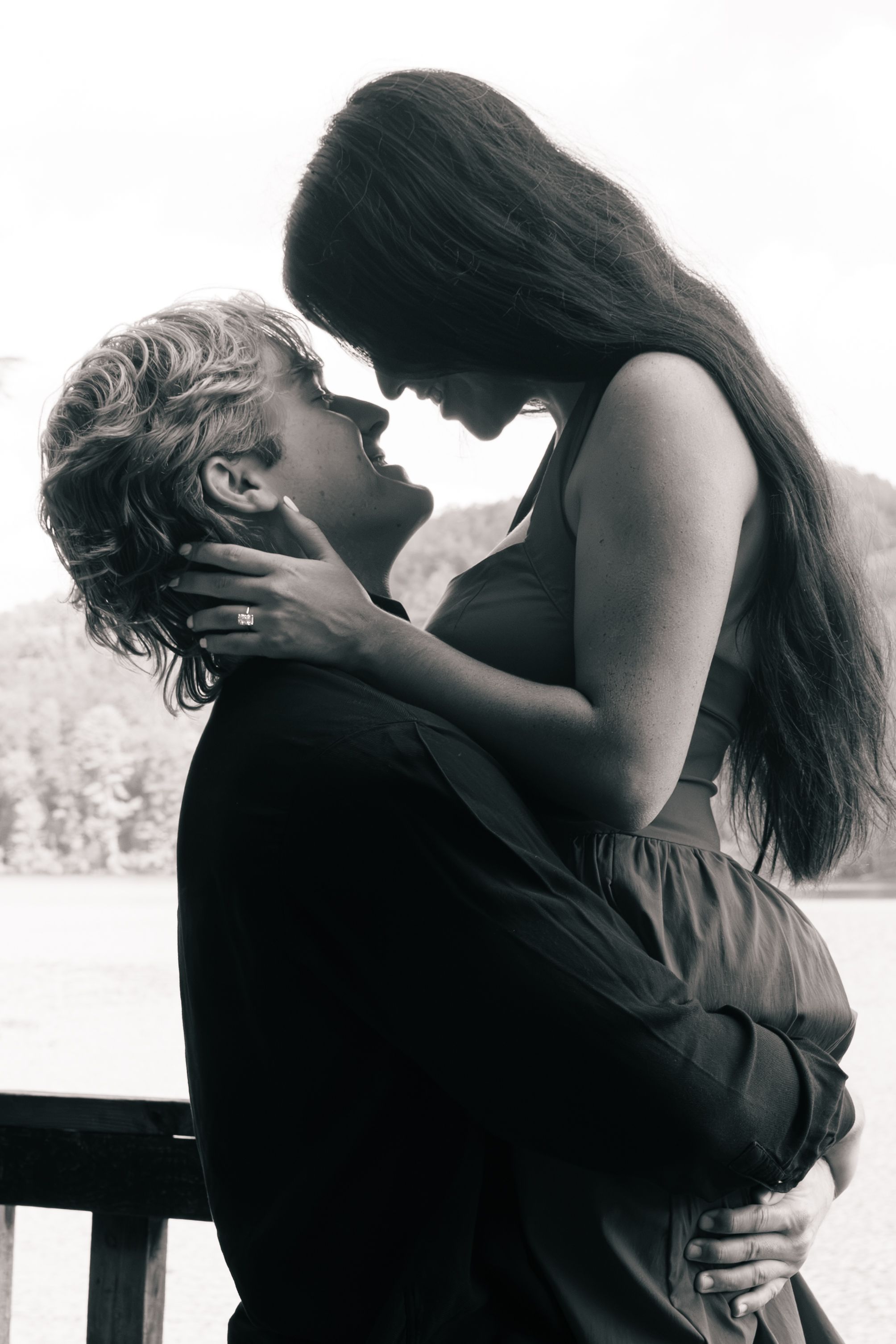 Black and white photo of a romantic couple embracing and about to kiss on a wooden deck beside a calm lake with forested hills behind.