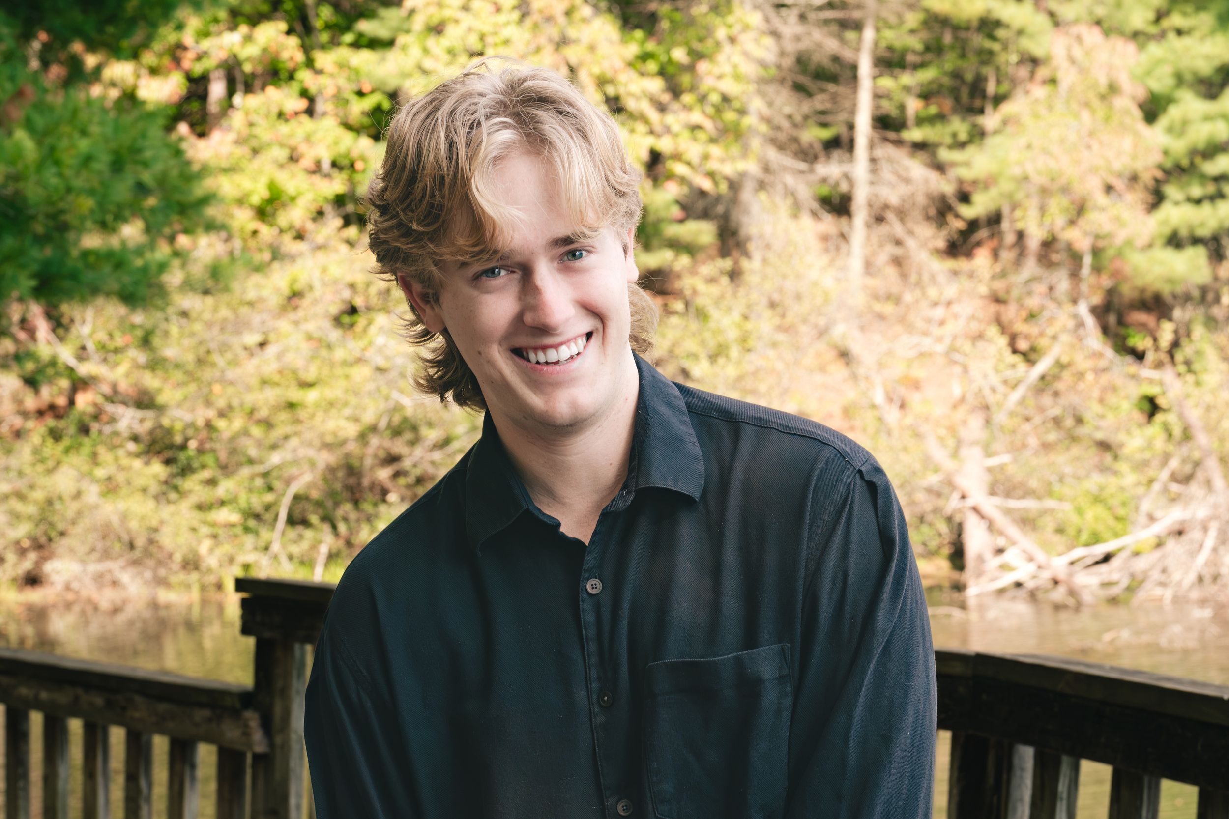 Smiling young man with wavy blond hair in a dark button-up shirt stands by a wooden railing with a wooded hillside and water behind him.