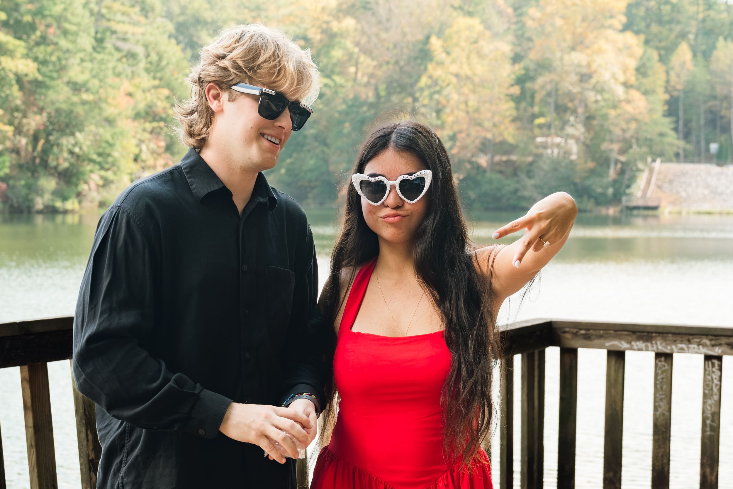 Man in black shirt and sunglasses stands beside a woman in a red dress and heart-shaped sunglasses on a lakeside wooden deck.
