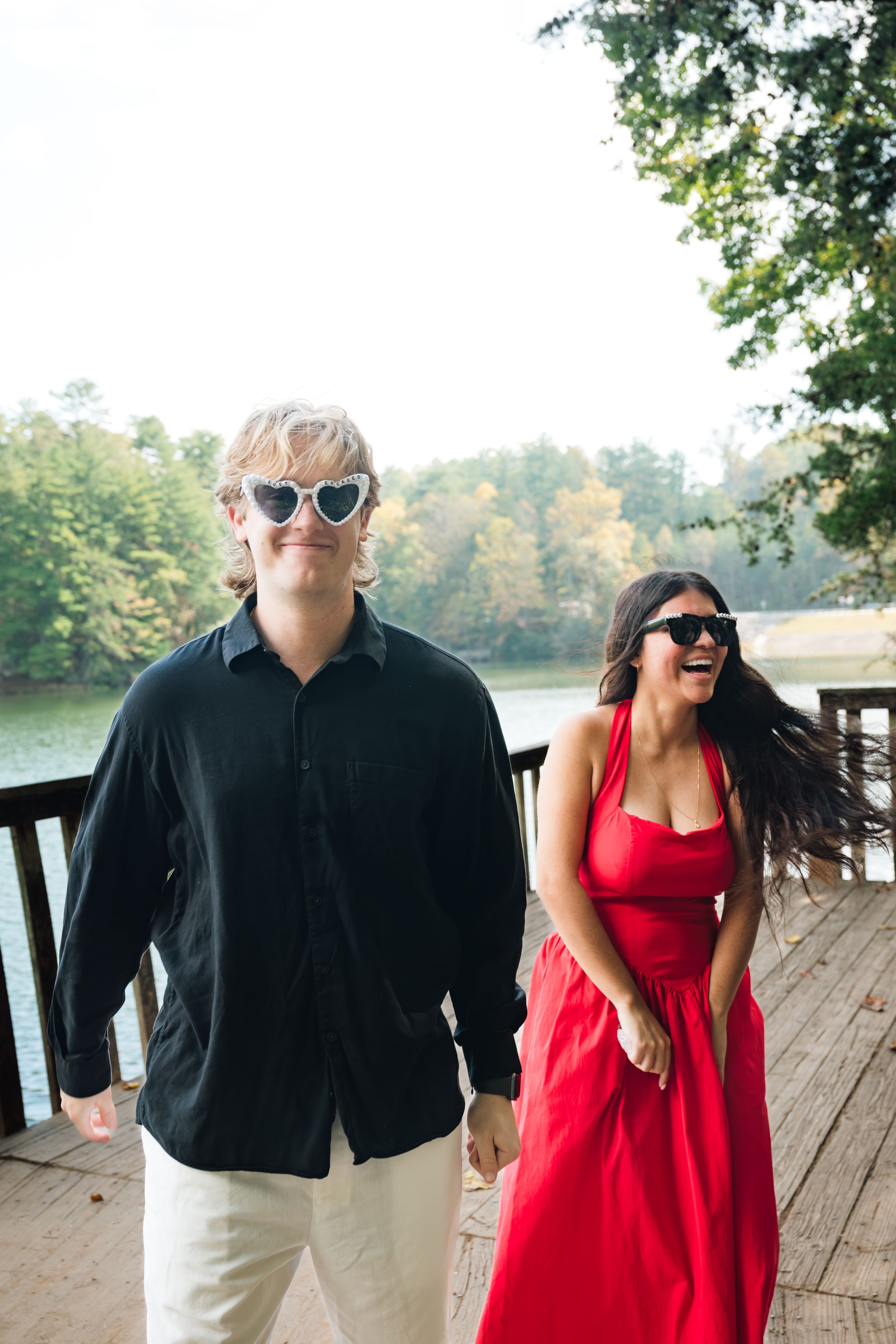 A laughing man in white heart-shaped sunglasses and a woman in a red dress walk along a wooden lakeside deck surrounded by trees.