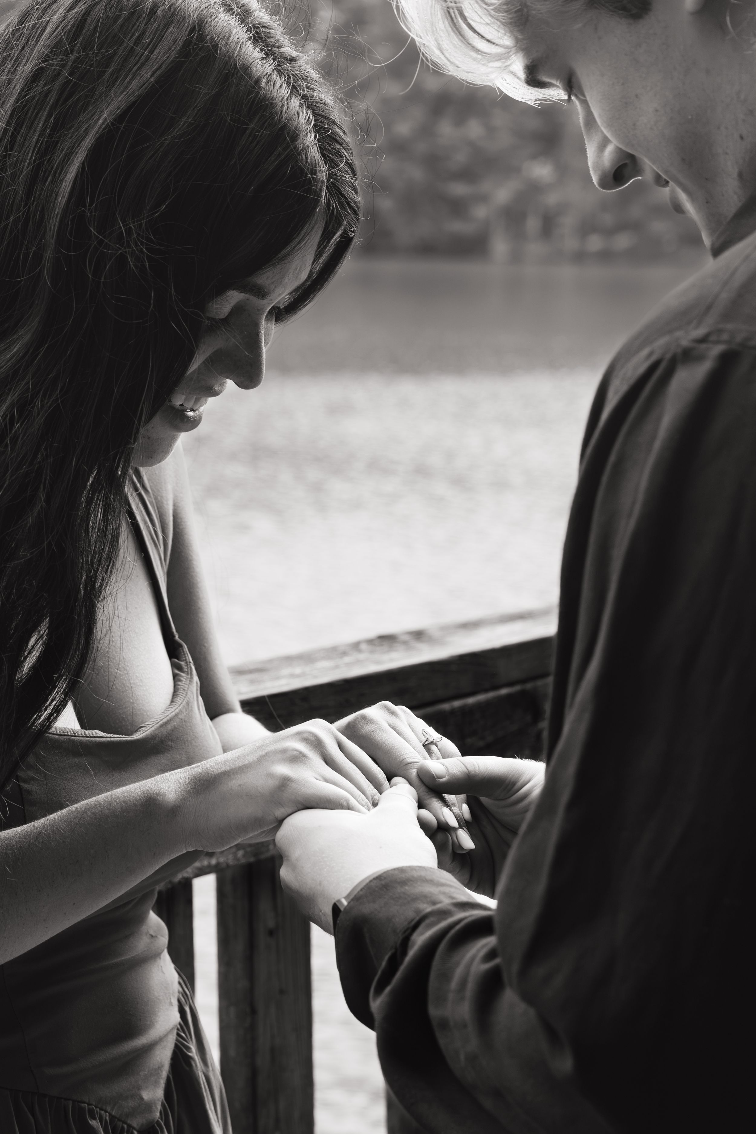 Black and white photo of a smiling woman and a man holding hands as he slides a ring onto her finger beside a calm lake.