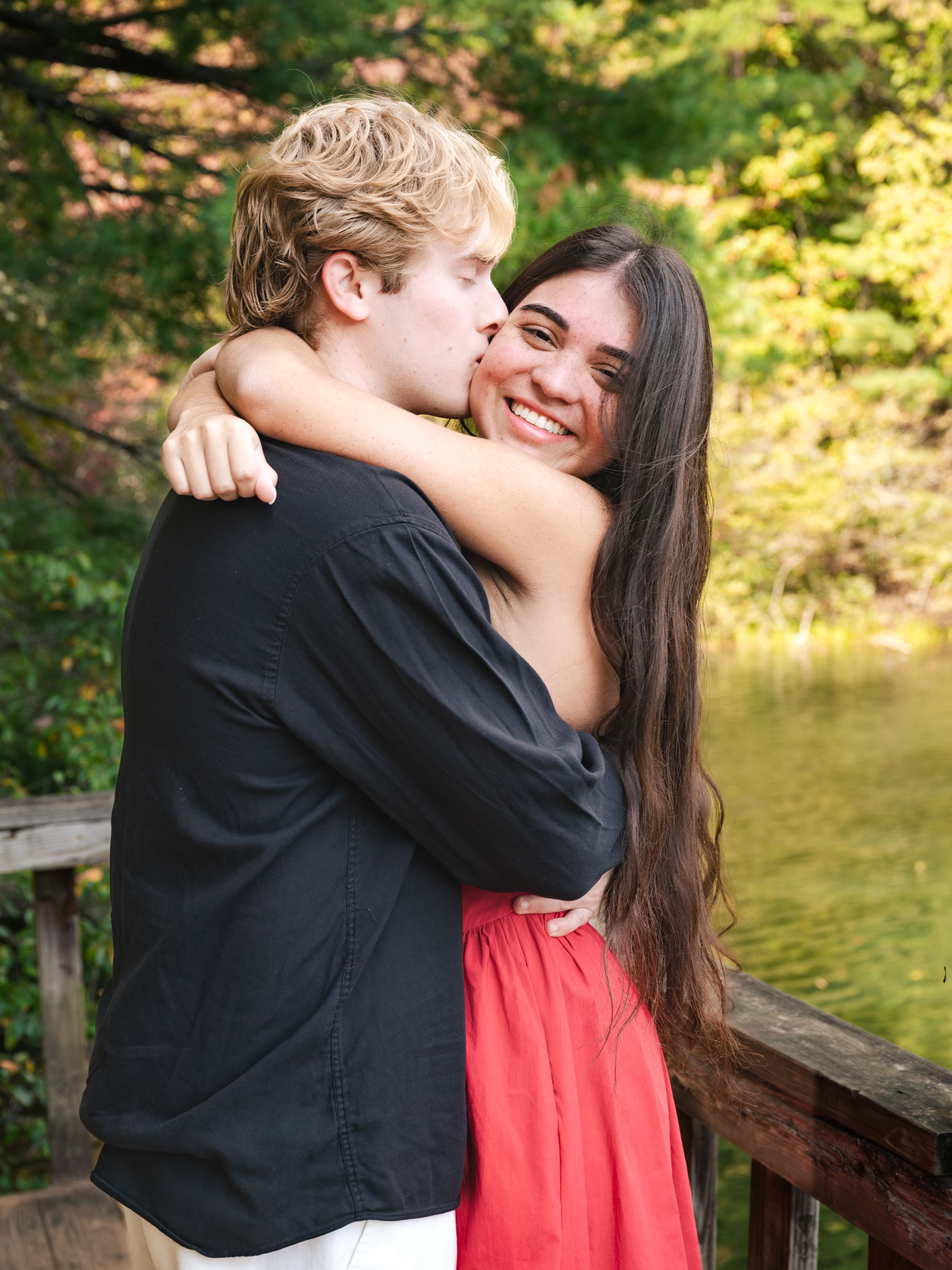 A couple embraces on a wooden dock by a calm lake as the man kisses the smiling woman's cheek, surrounded by green autumn trees.