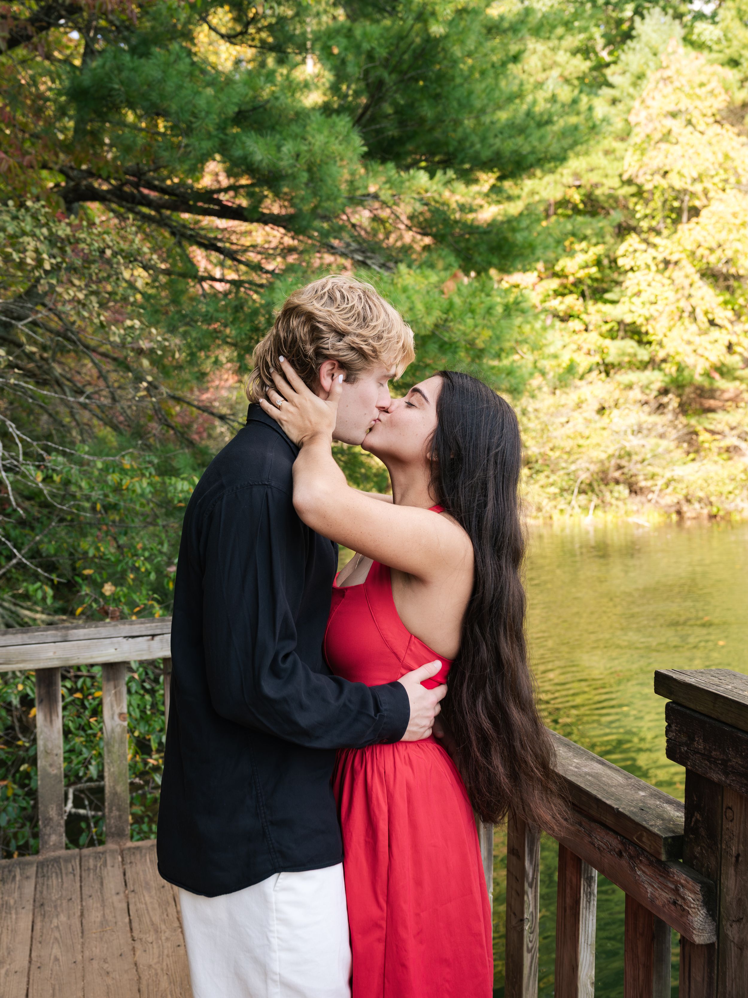 Couple kissing on a wooden dock by a calm lake, he wears a black shirt, she a red dress, with autumn trees behind.