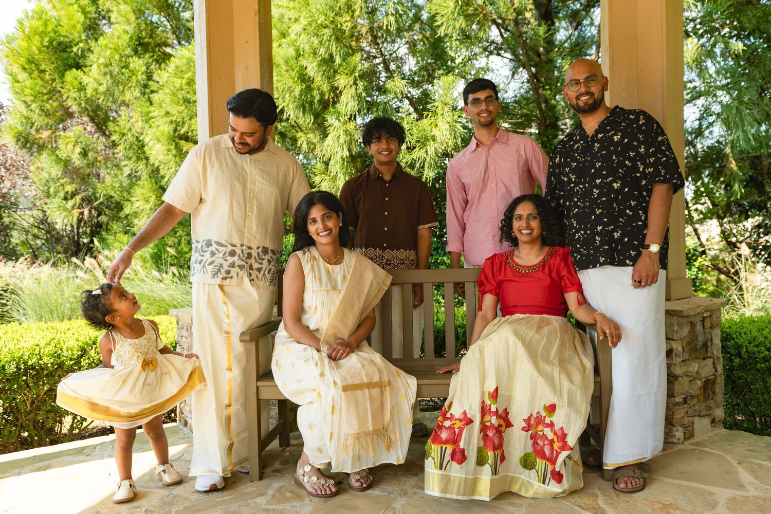 Family of seven in traditional cream and colorful outfits poses on a wooden bench and patio in a sunny garden, with a toddler in a yellow dr