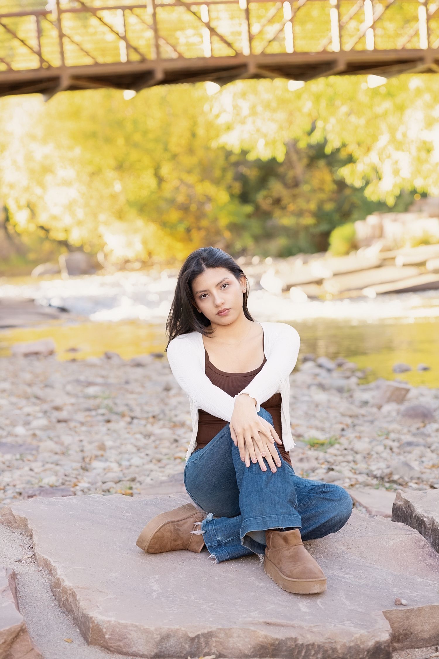 Young woman in white cardigan and jeans sitting on rocks near a creek with a wooden bridge backdrop.