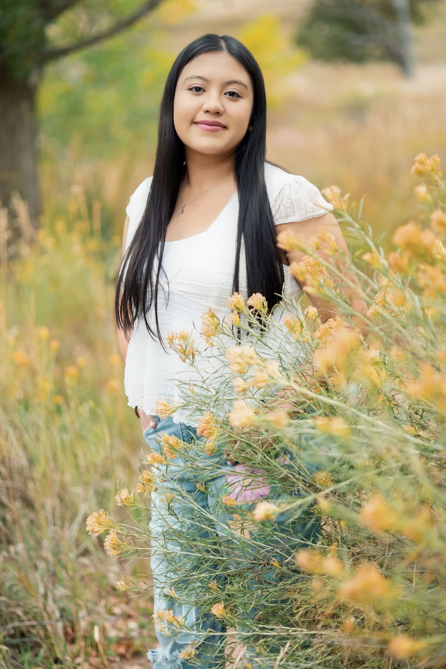 Young woman with long black hair in white top stands among yellow wildflowers in an autumn field.