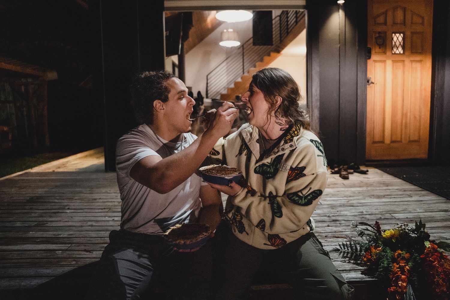 Couple sharing a playful moment on a wooden porch at night, one feeding the other, with a warm glowing doorway and colorful flowers nearby.